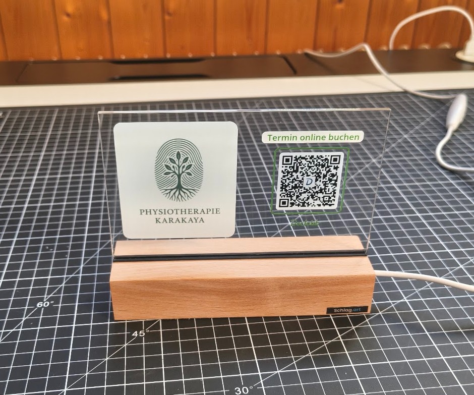 Kundenbeispiel: Acryl-Aufsteller mit Holzfuß, Logo Physiotherapie Karakaya und QR-Code. Logo und Link kommen vom Kunden.