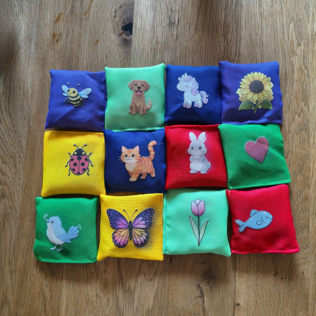 Zwölf kleine Stoffkissen auf einem Holzboden. UV-Druck mit den Motiven Biene, Hund, Einhorn, Sonnenblume, Marienkäfer, Katze, Hase, Herz, Vogel, Schmetterling, Tulpe und Fisch.